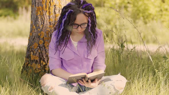 A Woman in a Purple Shirt and Glasses Reads a Literature Under a Tree