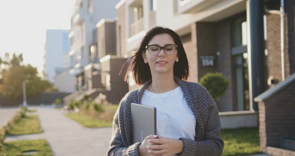 Woman Walking Through of Urban Street, Holding Laptop Device at Windy Spring Weather at Sunset