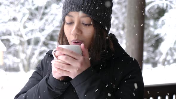 Beautiful woman holding cup of tea and warming hands on cold winter day during snowfall