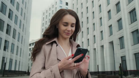 Cheerful young Caucasian woman standing in city street and texting on mobile phone with smile.