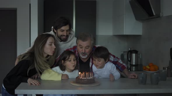 Happy Grandfather Blowing Candles on Birthday Cake Surrounded By His Family