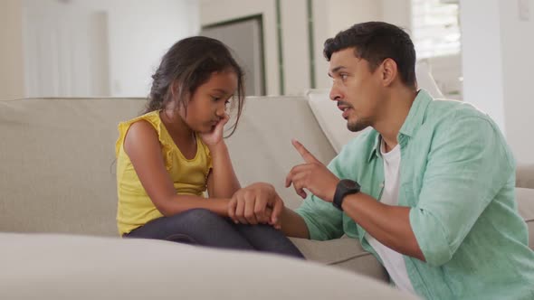 Hispanic father telling off his young upset daughter sitting on sofa