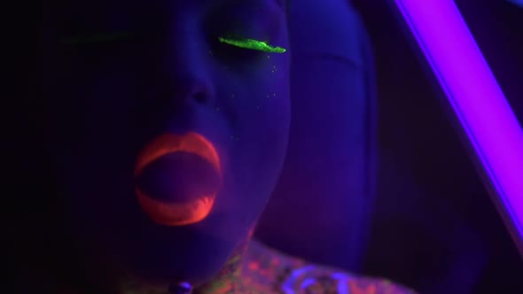 Portrait of a Young Beautiful Woman with Fluorescent Makeup Inhaling Hookah Under UV Black Light