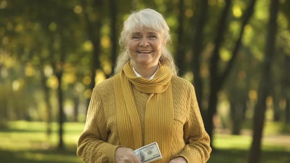 Smiling Old Woman Presenting Stack of Dollar Bills in Park, Planned Retirement