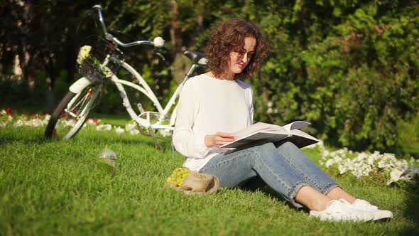 Attractive Woman in White Shirt and Blue Jeans is Reading a Book Sitting on the Grass in Park During