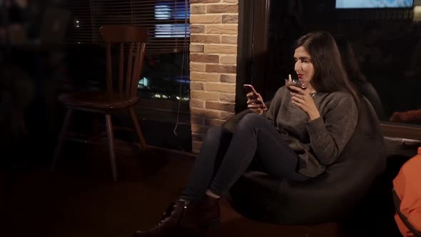 Cheerful Girl with Smartphone in Hands Is Drinking Cocktail From Goblet in Cafe