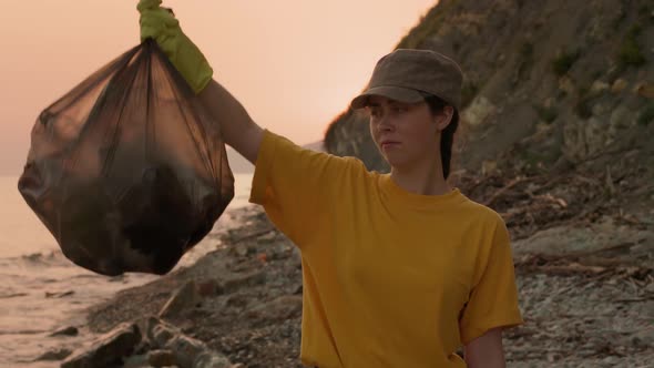 Woman in rubber gloves picks up a plastic bag full of garbage and expresses dissatisfaction.