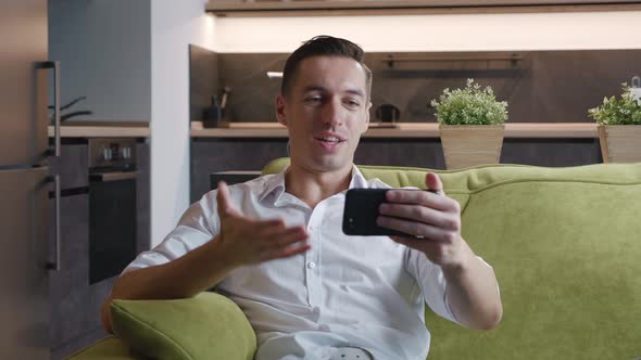 Smiling Young Man with Smartphone Making Video Call While Sitting on Couch in Living Room