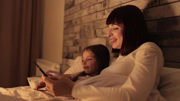 Pretty Likable Mother Lying on Bed Under the Blanket with Little Daughter Using Digital Tablet