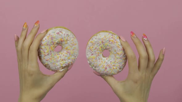 Womens Hands Holding Two White, Delicious, Sprinkled Donuts on Pink Background.