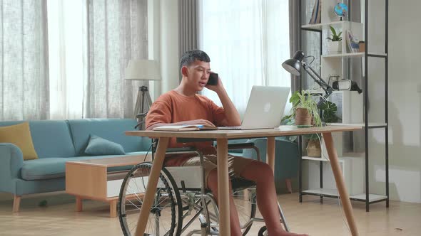 Asian Boy Sitting In A Wheelchair While Using Laptop Computer And Talking On Mobile Phone At Home