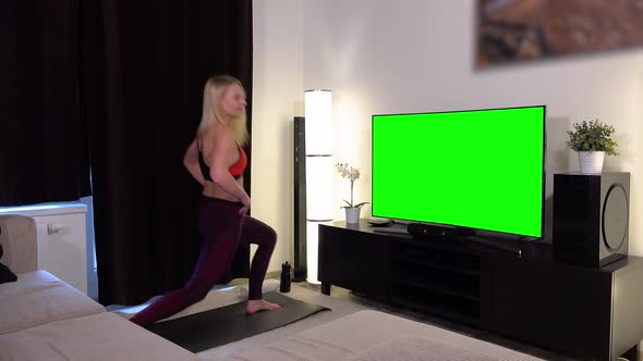 A young beautiful woman does lunges in front of a TV with a green screen in an apartment