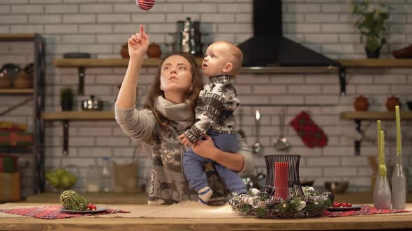 Pretty Young Woman Lifts Up the Baby in Her Arms Standing in Modern Kitchen Showing To Son Bright