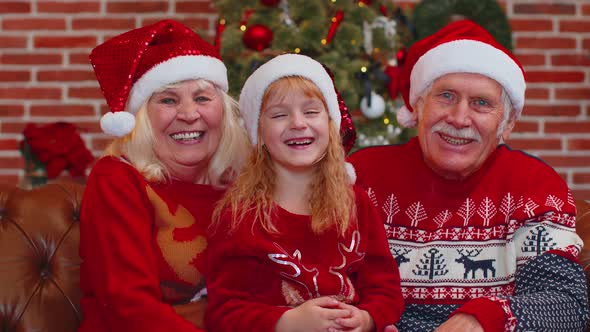Senior Grandparents with Granddaughter in Santa Hats Laughing Out Loud at Home Near Christmas Tree