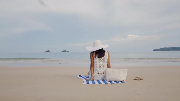 Woman In Sunhat Sitting On Beach Towel Looking Out To Sea