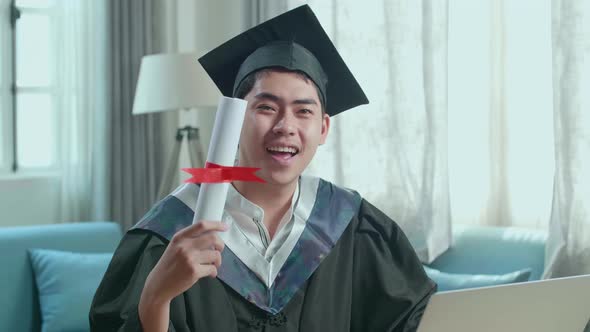 Excited Asian Man Smiling And Showing Off A University Certificate To Camera At Home