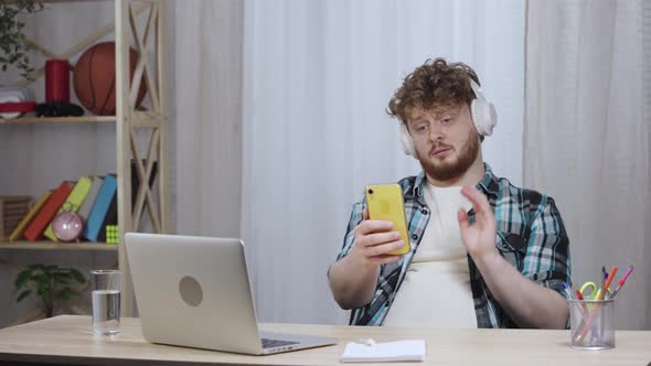 Young Man in Checkered Shirt Typing on Laptop Keyboard in Big White Headphones Then Talking on Video