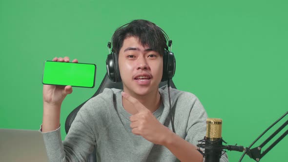 Smiling Asian Man With Headphone Reviewing Green Screen Mobile Phone On Green Screen