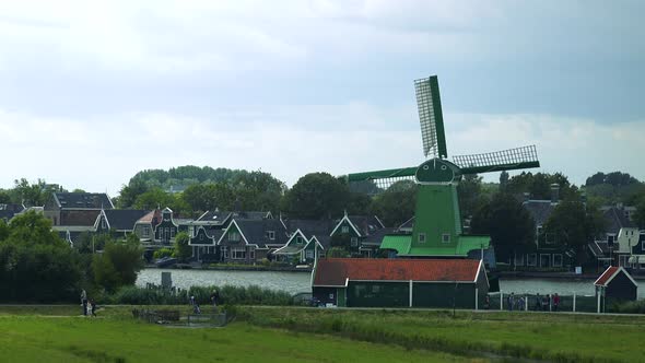 A Green Windmill By a River in a Rural Area, Its Vane Spins in the Wind, Buildings, Trees and Sky