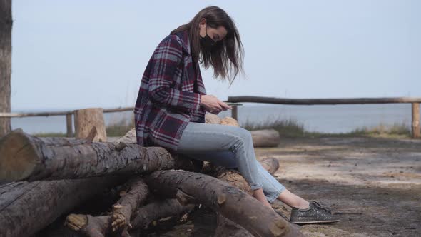 Side View of Brunette Girl in Face Mask Using Phone As Sitting on Log Next To the River. Young