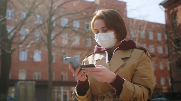 Happy Woman in Medical Mask Outdoor Holding Credit Card and Smartphone Buying Online Internet.