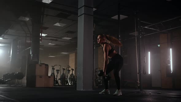 A Strong Woman Makes Efforts and Overcoming Difficulties Lifts a Dumbbell in a Dark Gym