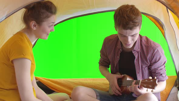 Young Man Plays Ukulele in Tent with Woman Back Wall is Chromakey