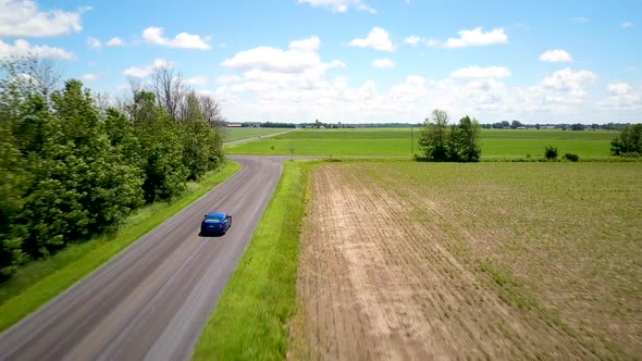 Aerial drone footage following an electric blue car driving up to an intersection on a country road