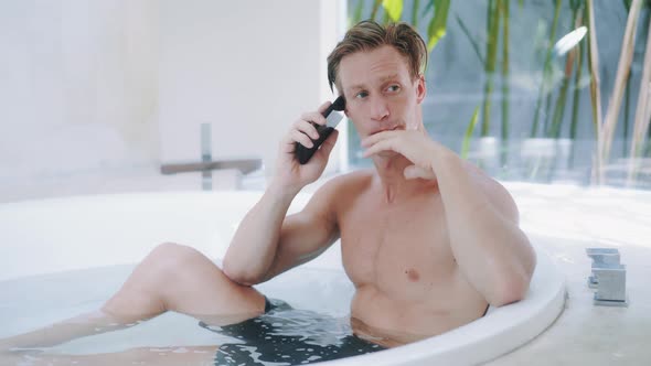 Muscular Blond Man Talks on Smartphone Sitting in Bathtub at Luxury Apartment