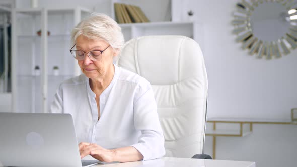 Concentrated aged lady manager in glasses types on grey laptop keyboard