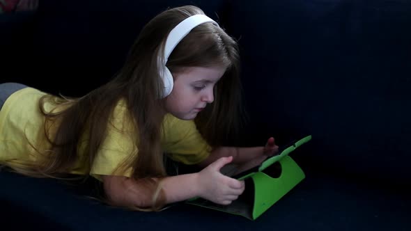 Cute small kid girl in headphones using tablet lying on sofa