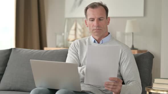 Middle Aged Businessman Doing Paperwork with Laptop on Sofa