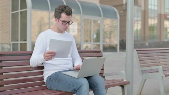 Young Man Reacting to Loss While Reading Documents and Working on Laptop Outdoor