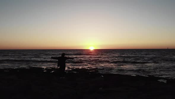 Black Silhouette of Woman on Rocky Sea Beach Hands Spread Wite During Sunset Over Sea with Waves