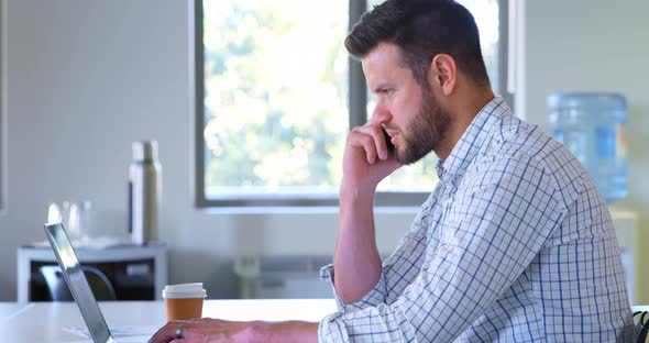 Side view of young caucasian male executive talking on mobile phone while using laptop in modern off