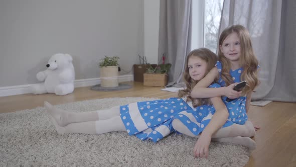 Two Cute Caucasian Girls Sitting on Soft Carpet Indoors with Remote Control, Twin Sisters in Blue