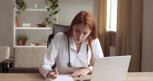 Portrait of Young Nurse Red-haired Young Woman Doctor Sits in Office in Clinic Looks at Laptop