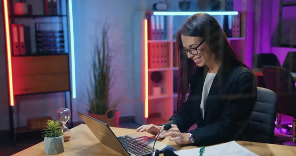 Brunette in Stylish wear Sitting at Her Workplace in Evening Office and Typing on Laptop