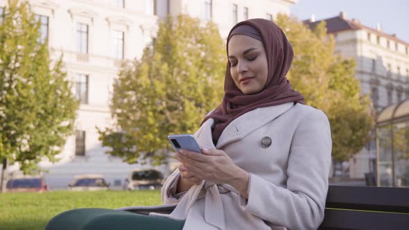 A Young Beautiful Muslim Woman Works on a Smartphone with a Smile As She Sits in a Park