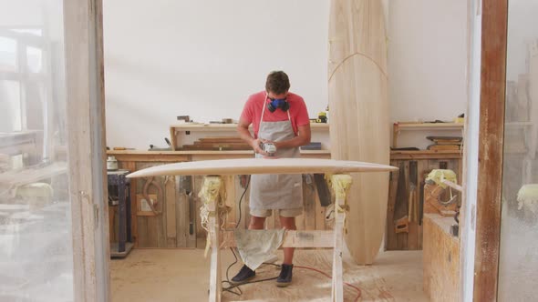 Caucasian male surfboard maker wearing a breathing face mask and shaping a wooden surfboard