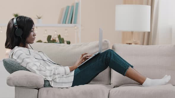 Young Female Freelancer Student Lying on Sofa at Home Typing on Laptop Listening to Music on