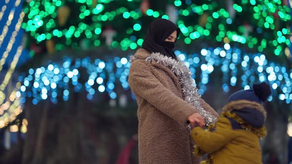 Carefree Excited Middle Eastern Mother and Son Spinning in City with Christmas Lights at Background