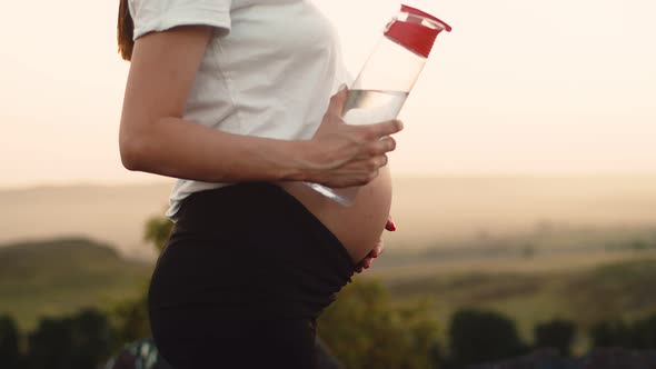 Closeup Stomach of Pregnant Woman Holding Hand on Stomach and Holding Bottle of Water Side View