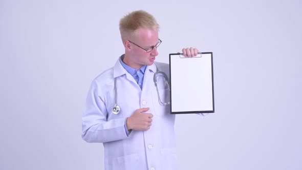 Serious Young Blonde Man Doctor Showing Clipboard and Giving Thumbs Down