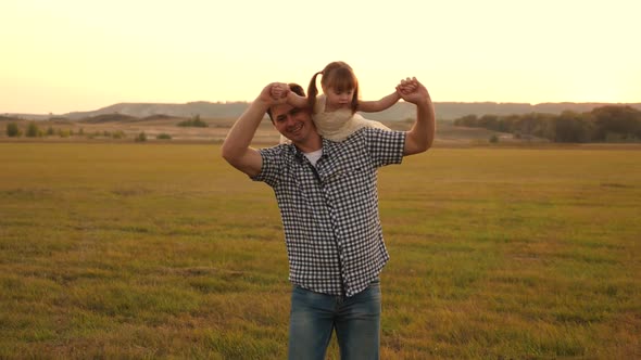 Happy Family Resting in the Park. Father Walks with His Daughter on His Shoulders in Rays of Sunset