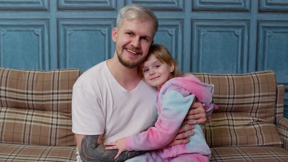 Father and Little Child Daughter Kid in Pajamas Sit on Couch in Room Smiling Looking at Camera