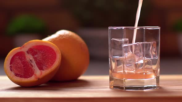 Fresh Grapefruit Juice Pouring Into a Transparent Glass with Ice. Sliced Grapefruits on a Table