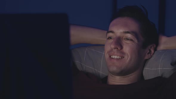 Smiling Young Man Watching a Movie on Laptop While Resting on Bed at Night