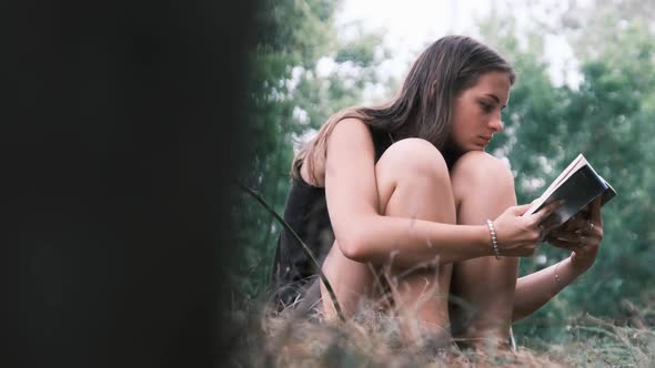 Young Woman Reads a Book Sitting on the Grass in Nature at Summer Day