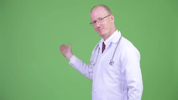 Happy Mature Bald Man Doctor Showing Something Against Green Background